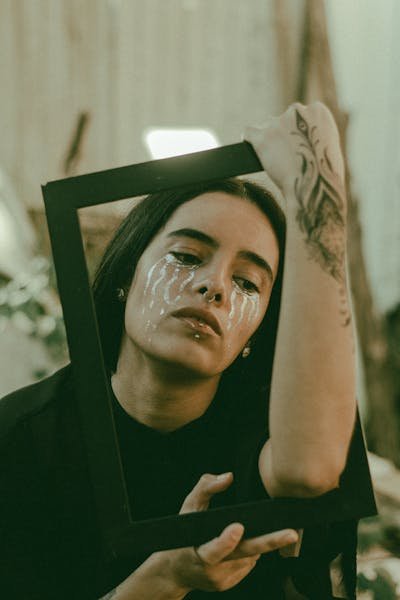 A creative portrait featuring a woman holding a frame with artistic makeup and tattoos.