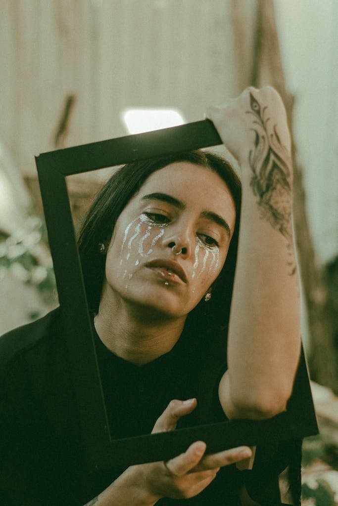 A creative portrait featuring a woman holding a frame with artistic makeup and tattoos.