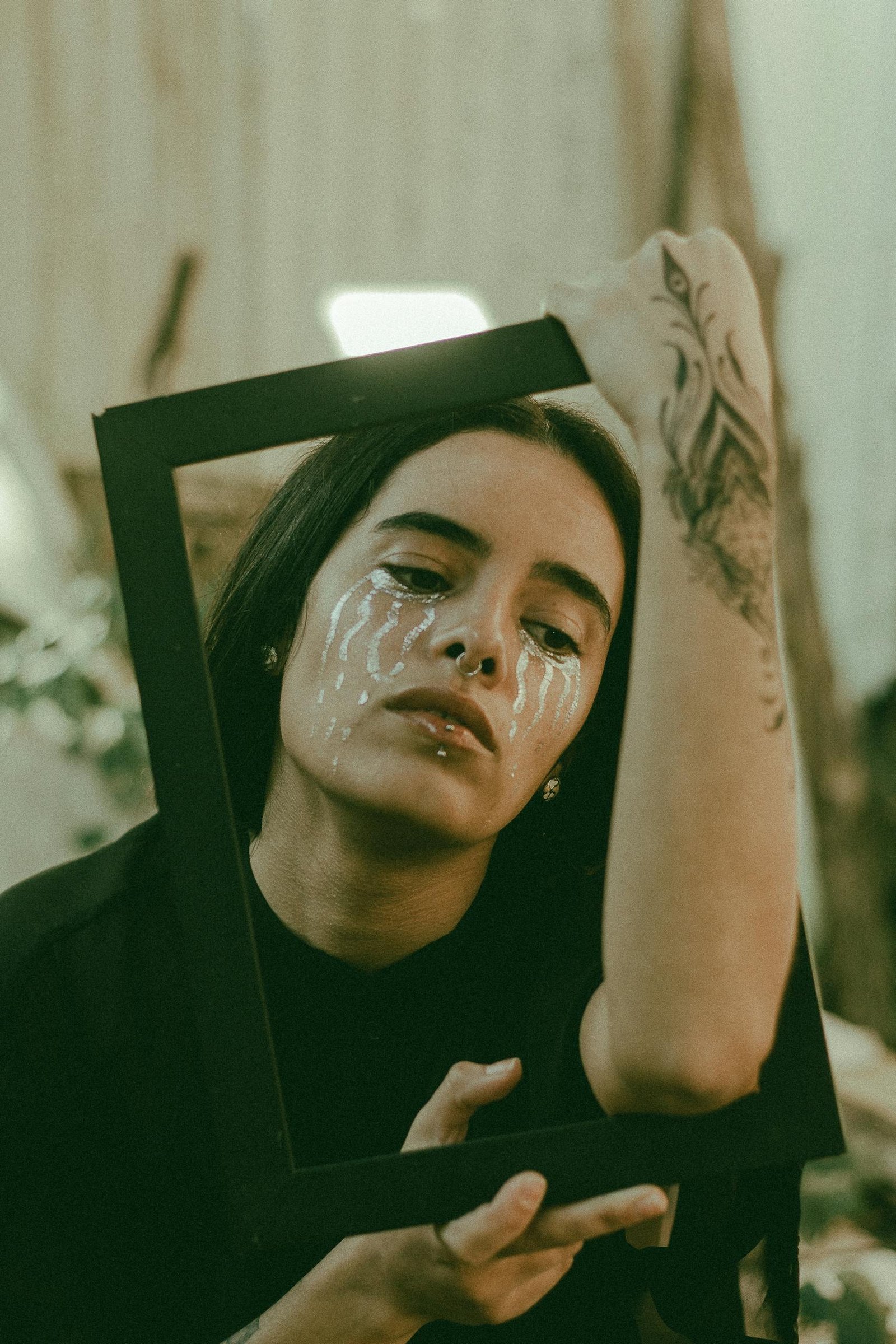 A creative portrait featuring a woman holding a frame with artistic makeup and tattoos.