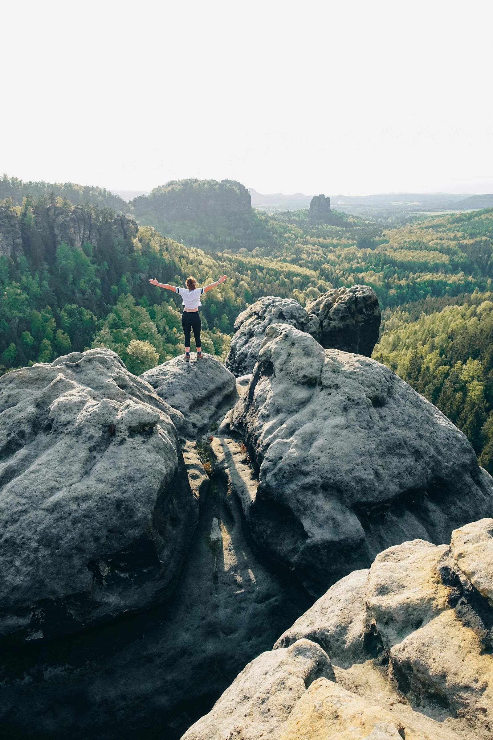 Woman standing on Elbe Sandstone Mountains, arms raised, surrounded by stunning natural scenery.