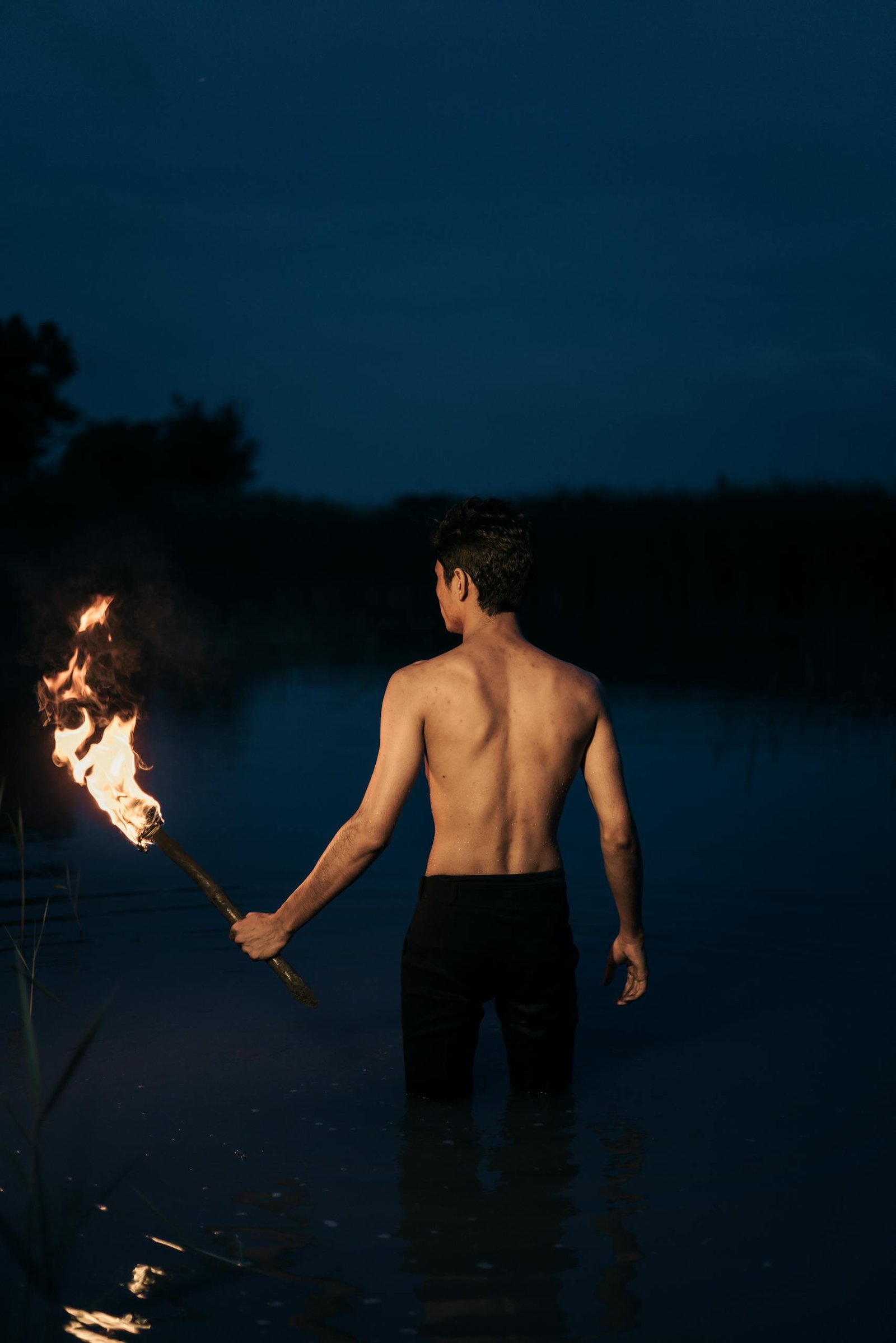 A topless man holds a fiery torch at night in calm water, creating a dramatic silhouette.